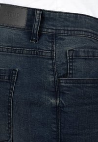 Tmavě modré džínové kalhoty s viditelným šitím, zadními kapsami, poutky na opasek a černou etiketou s slabým textem na pasu.