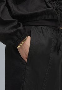 Lettet sort stof outfit med elastiske manchetter og talje, samt en synlig lomme. Et guld armbånd bæres på håndleddet.