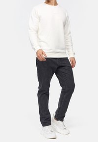 Sudadera blanca con cuello redondo, mangas acanaladas, combinada con jeans gris oscuro y zapatillas blancas, presenta una textura suave y un branding mínimo.