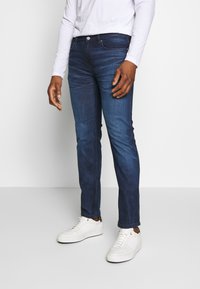 Jeans de mezclilla azul oscuro con corte ajustado, que presentan un clásico diseño de cinco bolsillos y un ligero desvanecimiento. Combinados con zapatillas blancas.