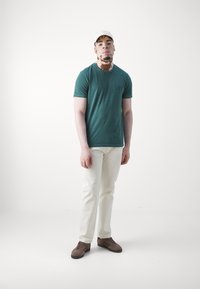 Grønn bomulls t-skjorte med rund hals og subtilt logo, kombinert med hvite jeans og brune joggesko. Minimalistisk design, ensfarget bakgrunn.