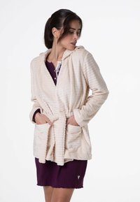 Robe con cappuccio di colore crema, con texture, dotata di cintura, due tasche e dettagli a costine; indossata sopra un indumento di colore viola scuro con colletto in pizzo.