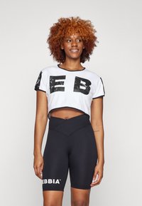 Hvid cropped t-shirt med store sorte blokbogstaver og sorte detaljer, parret med højtaljede sorte shorts med hvid branding.