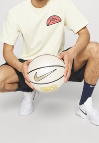 Bledě žluté tričko s růžovou grafikou basketbalového míče, černé šortky, držící bílý basketbal Nike Versa Tack se zlatými akcenty, obuté v bílých botách.