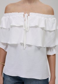 Witte off-shoulder top met meerdere laagjes ruches, trekkoorddetails bij de halslijn en een getextureerde stof. Gecombineerd met denim.
