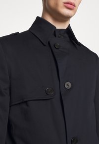 Marinblå trenchcoat med klassisk krage, dubbelknappstockning och slät tygkänsla. Detaljerad sömnad på slaget.