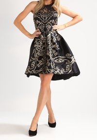 Czarna satynowa sukienka z srebrnymi haftami, w kształcie litery A, bez rękawów, z wycięciem przy dekolcie. Czarne szpilki dopełniają look.