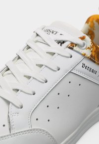 Λευκό δερμάτινο αθλητικό παπούτσι με λευκά κορδόνια, διατρητική πλαϊνή επιφάνεια, χρυσό μοτίβο στον γιακά και μαύρο λογότυπο "Versace" στο πλάι.