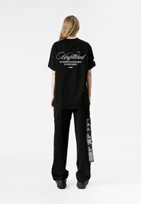 Camiseta negra de gran tamaño con texto impreso en blanco en la parte posterior. Combinada con pantalones sueltos negros, también adornados con texto. Calzado casual.