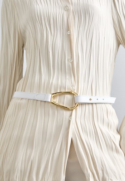 Witte leren riem met een grote gouden ovale gesp, gedragen over een crèmekleurig, geplooid overhemd met knopen. Soepele textuur met minimale hardwaredetails.