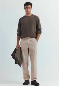 Beige op maat gemaakte broek gecombineerd met een donker geribbelde trui, met een bijpassende trui in de hand, afgewerkt met donkere schoenen; gladde textuur, neutrale kleuren.