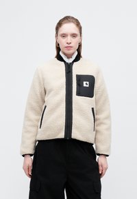 Beige fleece-takki, jossa on mustia yksityiskohtia, vetoketjulla varustettu etuosa ja rintatasku. Suora kaulus ja hihansuut, pehmeä pinta, suorakulmainen muoto.
