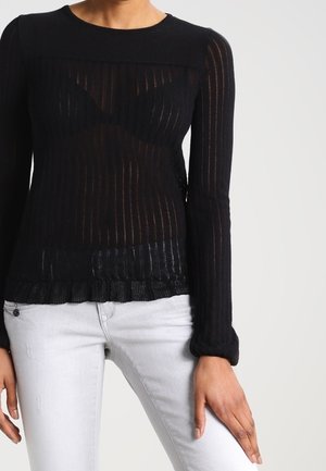 Femme portant un haut moulant en maille noire transparente à manches longues et un jean gris clair, debout avec une main posée sur la cuisse.