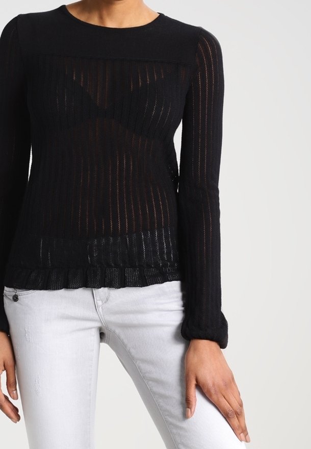 Femme portant un haut moulant en maille noire transparente à manches longues et un jean gris clair, debout avec une main posée sur la cuisse.