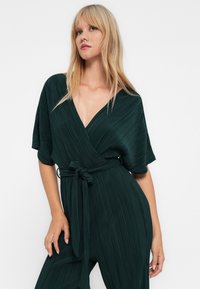 En blond kvinne iført en mørkegrønn plissédrapert jumpsuit med korte ermer og knyttet belte, poserer mot en hvit bakgrunn.