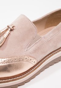 Mocassin en daim rose clair avec pompons métalliques en or rose, bordure perforée et semelle épaisse en caoutchouc avec surpiqûres blanches.
