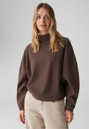 Femme aux longs cheveux blonds portant un pull marron oversized et un pantalon beige clair, se tenant devant un fond gris uni.