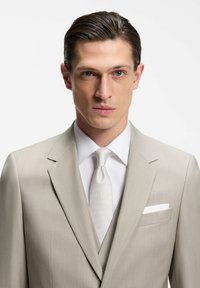 Homme aux cheveux lissés en arrière, portant un costume beige clair, une chemise blanche, une cravate assortie et un mouchoir blanc dans la poche, sur un fond uni.
