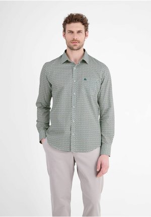 Man met kort krullend haar, die een lange mouwen patroon blouse en lichte broek draagt, staat met één hand in zijn zak.