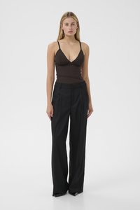 Donna che indossa un top senza maniche marrone scuro con spalline sottili e pantaloni larghi neri a vita alta, in piedi su uno sfondo bianco.