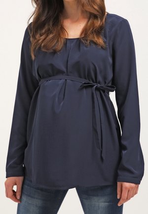 Mujer con blusa premamá de manga larga azul marino con lazada lateral y vaqueros azules, mostrando el torso y los brazos.