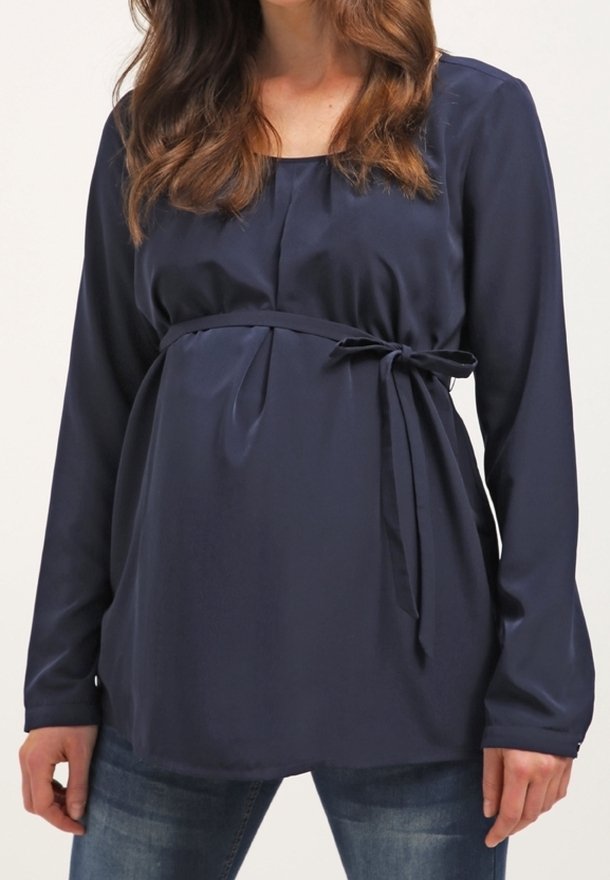 Femme portant un chemisier de maternité bleu marine à manches longues avec un lien latéral et un jean bleu, montrant le haut du corps et les bras.