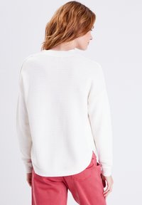 Maglione bianco a coste con scollo rotondo e spalle scese, abbinato a pantaloni rosa. Il maglione presenta un orlo leggermente curvo.
