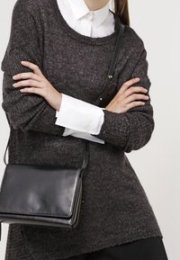 Tummanharmaa neulepaita leveällä kaula-aukolla, kerroksittain valkoisen kauluspaitaan päällä. Musta nahkainen olkalaukku, jossa kultainen vetoketjutalja.