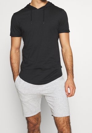 Mężczyzna w czarnej bluzie z krótkim rękawem i jasnoszarych krótkich spodenkach, stojący z jedną ręką w kieszeni na jednolitym tle.