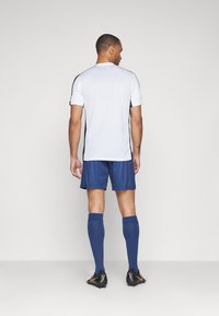 Vitt idrottströja med svarta detaljer, tillsammans med mörkblå shorts och knähöga blå strumpor. Svarta fotbollsskor med orange detaljer.