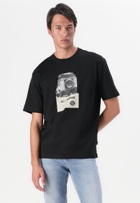 Camiseta de algodón negra con un estampado gráfico de un vehículo vintage y una etiqueta de texto sobre un fondo claro. Manga corta y un ajuste relajado.