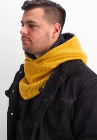 Knitted yellow cowl scarf with a textured pattern, worn over a black outerwear jacket featuring a shearling collar and visible buttons.