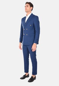 Abito doppiopetto blu navy con bottoni dorati, vestibilità su misura, pantaloni coordinati e mocassini neri, con una camicia bianca sotto.