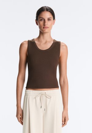 Žena s tamnom kosom vezanom unatrag, nosi smeđi rebrasti top bez rukava i svijetlo bež hlače s vezicama, stoji ispred jednobojne pozadine.