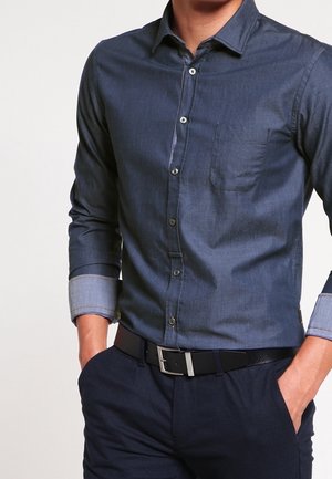 Homme portant une chemise bleu marine à boutons avec les manches retroussées, ceinture noire et pantalon sombre, mains dans les poches devant un fond uni.