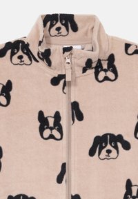 Veste en polaire beige avec un col haut, ornée d'un motif de visage de chien noir. Elle possède une fermeture éclair à l'avant et un tissu doux et texturé.