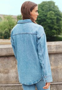 Lichtblauwe denimjas met een ontspannen pasvorm, spitse kraag en knoopmanchetten. Zichtbare stikseldetails en een licht gebogen onderrand.