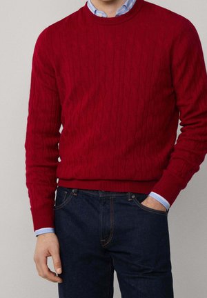 Homme portant un pull rouge en maille torsadée par-dessus une chemise bleue à col, accompagné d'un jean bleu foncé, une main dans la poche.