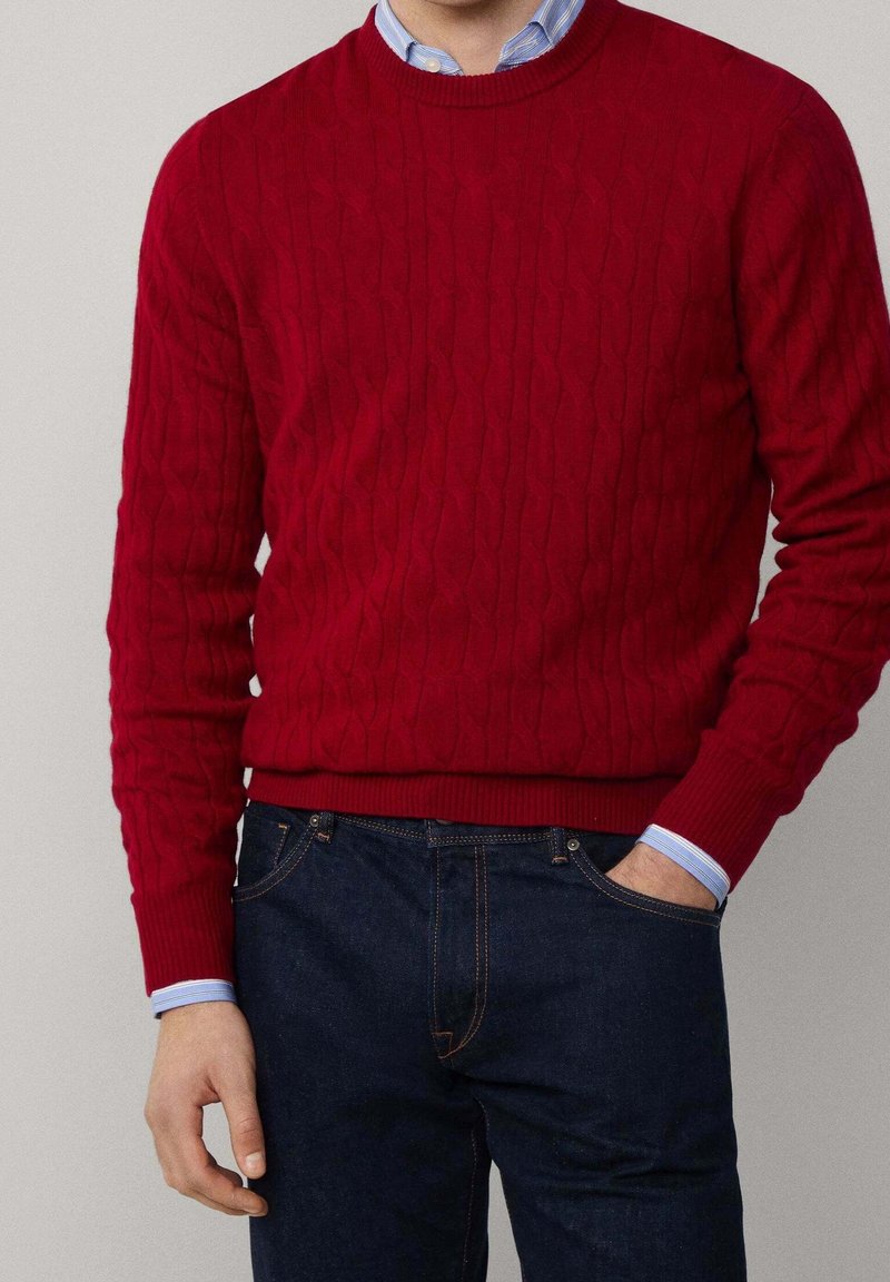 Homme portant un pull rouge en maille torsadée par-dessus une chemise bleue à col, accompagné d'un jean bleu foncé, une main dans la poche.