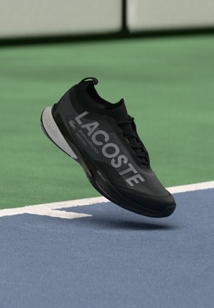 Chaussure de sport noire Lacoste flottant au-dessus d'une surface de court de tennis avec des couleurs vertes et bleues et des lignes blanches de délimitation.