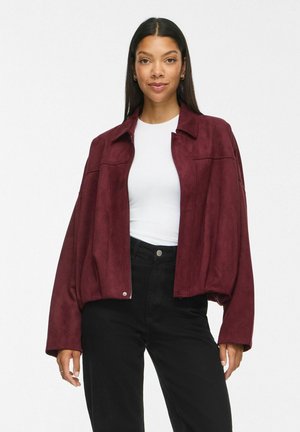 Femme aux longs cheveux raides et foncés, portant un haut blanc, une veste bordeaux ample et un pantalon noir taille haute, debout devant un fond uni.