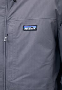 Veste imperméable grise avec une poche zippée, tissu texturé et un logo Patagonia mettant en avant des détails de montagne et de rayures colorées.