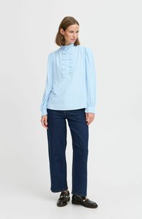 Helles blaues Blusenshirt mit hochgeschlossenem Kragen, gerafftem Frontdetail, langen Ärmeln und taillierten Manschetten, kombiniert mit dunklen Denim-Jeans und Slippern.