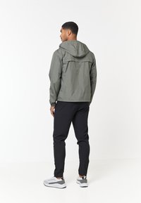 Letvægts, olivengrøn vandtæt jakke med hætte, elastiske manchetter og afslappet pasform, parret med sorte, snævre bukser og hvide sneakers.