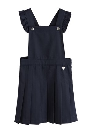 Young girl standing, wearing a white blouse with puffed sleeves and a black pleated pinafore dress with ruffled straps.