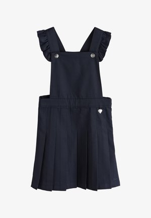 Ung pige står, iført en hvid bluse med puffede ærmer og en sort plisseret forklædekjole med flæsebesatte stropper.