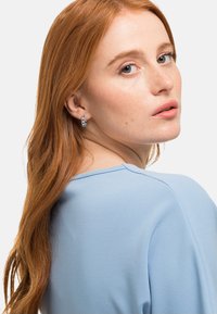 Top azzurro chiaro realizzato in tessuto morbido con vestibilità relax. Presenta un collo rotondo e dettagli delle cuciture sottili. Accento con piccoli orecchini ad anello in argento.