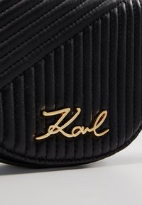 Surface en cuir matelassé noir avec des coutures diagonales et verticales et une signature métallique dorée "Karl" dans le coin inférieur droit.