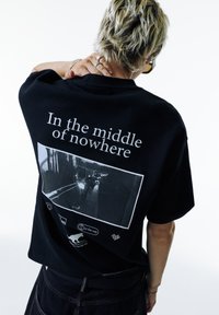 Persona che indossa una maglietta nera con la scritta bianca "In the middle of nowhere" e una foto di strada in bianco e nero sul retro, mentre posa con una mano sul collo.