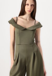 Olivengrønn jumpsuit med avtakbare ermer, tettsittende overdel, brede plisserte ben og myk, glatt tekstur.